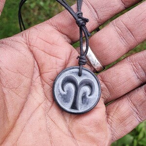 May include: A black cord necklace with a black and gray round pendant featuring the astrological symbol for Aries.