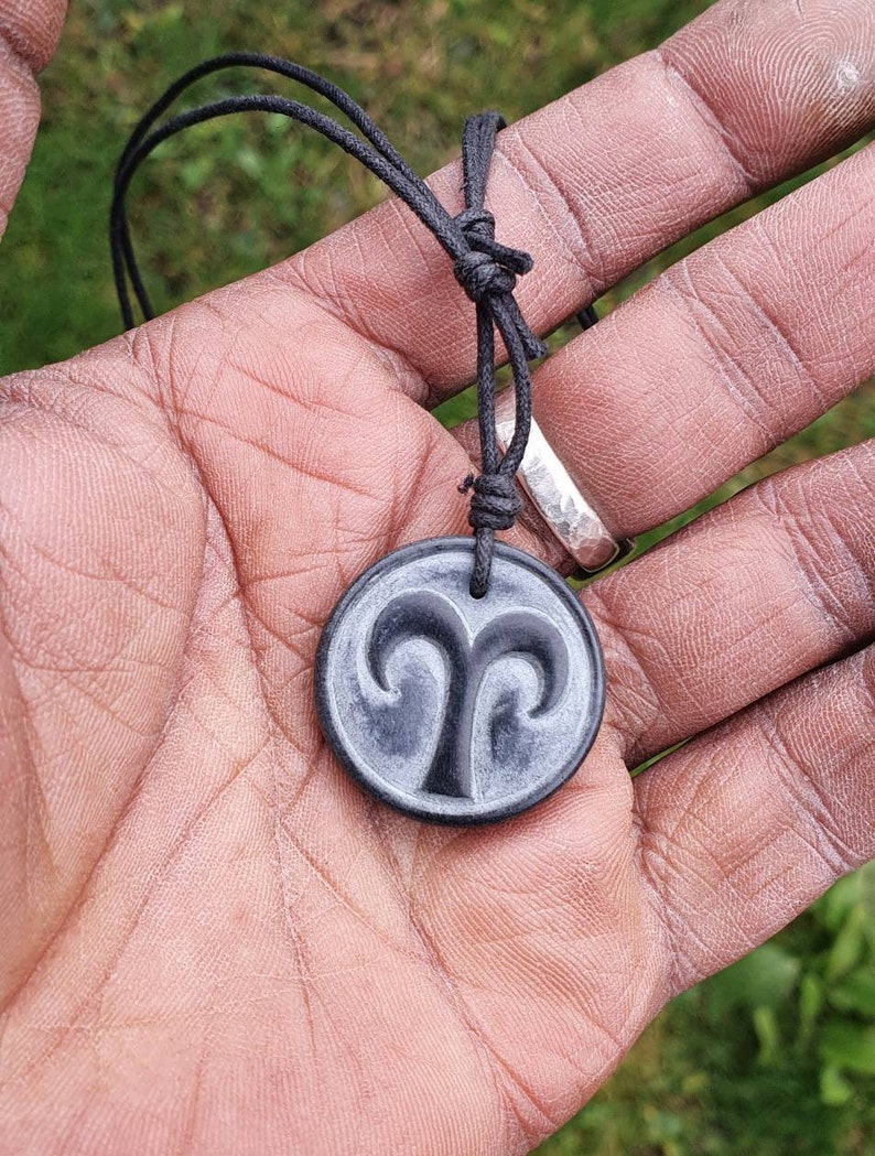 May include: A black cord necklace with a black and gray round pendant featuring the astrological symbol for Aries.