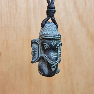 Pode incluir: Um pingente de madeira esculpida em preto e cinza da divindade hindu Ganesha, pendurado em um cord&atilde;o preto.
