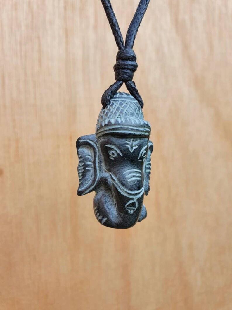 Pode incluir: Um pingente de madeira esculpida em preto e cinza da divindade hindu Ganesha, pendurado em um cord&atilde;o preto.