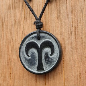 May include: A black necklace with a round pendant featuring a carved Aries zodiac symbol. The pendant is made of black stone with a silver-grey swirl pattern.