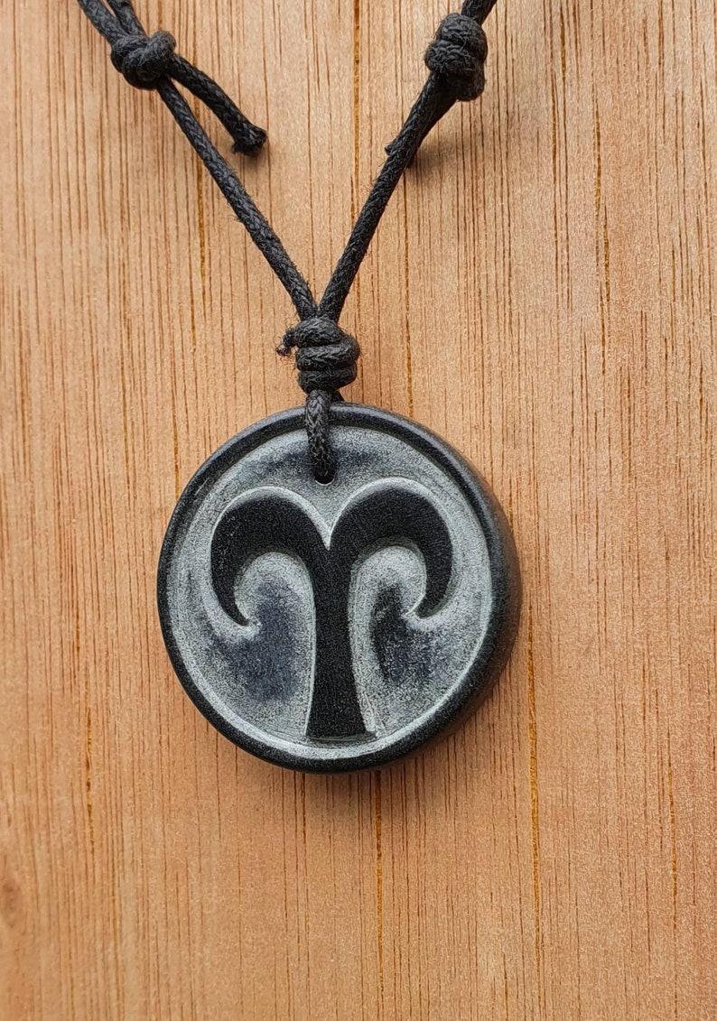 May include: A black necklace with a round pendant featuring a carved Aries zodiac symbol. The pendant is made of black stone with a silver-grey swirl pattern.