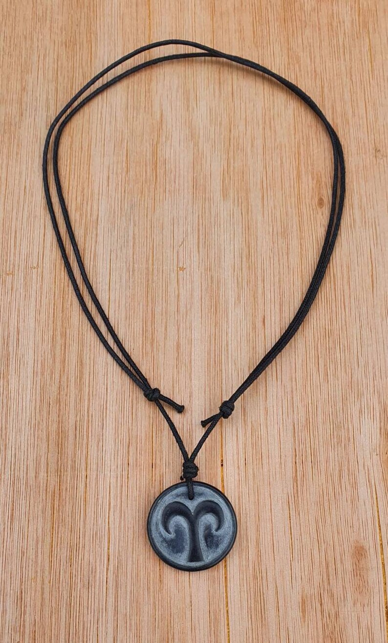 May include: A black cord necklace with a round gray pendant featuring the astrological symbol for Aries.