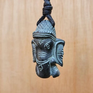 Pode incluir: Um pingente de pedra preta e verde que representa a divindade hindu Ganesha, o deus com cabe&ccedil;a de elefante dos come&ccedil;os, dos obst&aacute;culos e da sabedoria. O pingente est&aacute; pendurado em um cord&atilde;o preto.