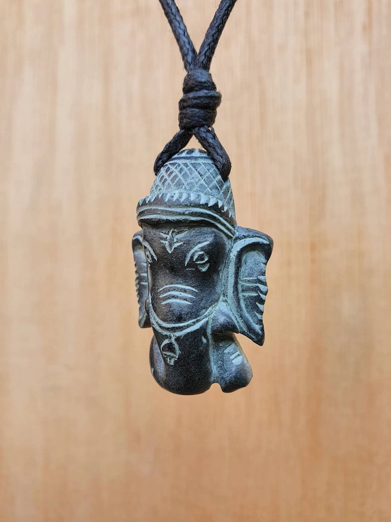 Pode incluir: Um pingente de pedra preta e verde que representa a divindade hindu Ganesha, o deus com cabe&ccedil;a de elefante dos come&ccedil;os, dos obst&aacute;culos e da sabedoria. O pingente est&aacute; pendurado em um cord&atilde;o preto.