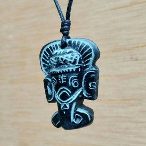 May include: Black pendant necklace featuring a carved wooden figure of a deity with intricate details. The figure has a large head with a headdress and a detailed face.