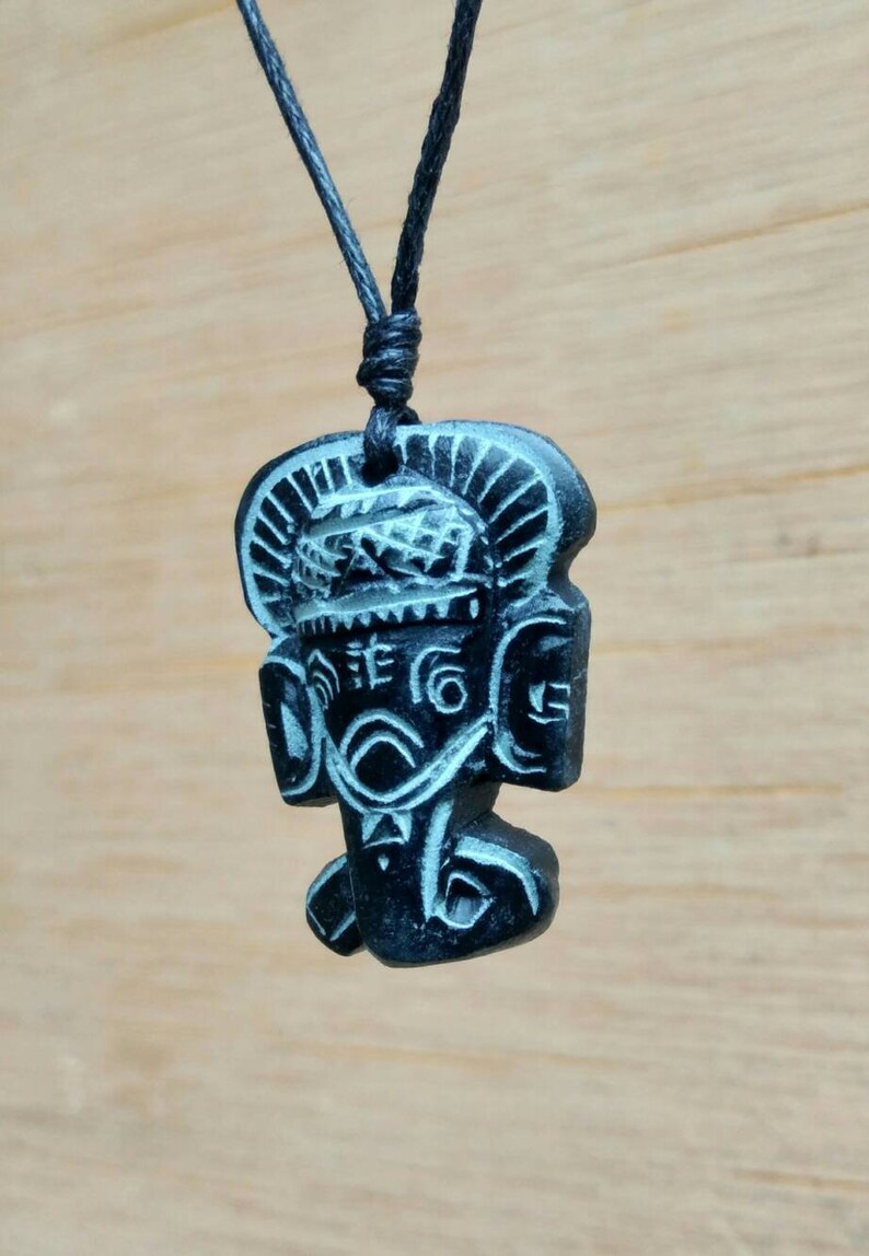 May include: Black pendant necklace featuring a carved wooden figure of a deity with intricate details. The figure has a large head with a headdress and a detailed face.