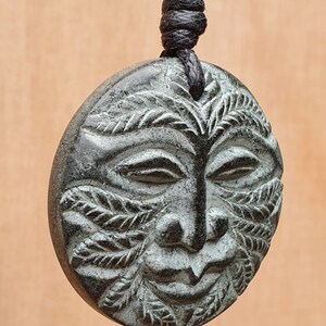 以下が含まれることがあります： 黒い紐に、顔と葉が彫刻された緑色の石のペンダントがぶら下がっている