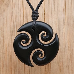 Può includere: Collana con ciondolo in legno nero di forma circolare con tre spirali intagliate. La collana è su un cordoncino nero.