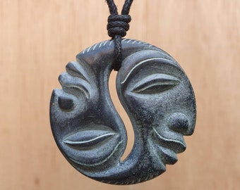 May include: A black necklace with a carved wooden pendant featuring two stylized faces, one with closed eyes and the other with open eyes, joined together in a yin-yang style.