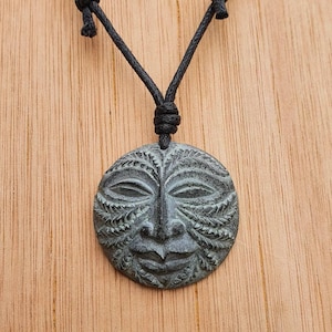May include: A round, green pendant with a carved face design hangs from a black cord necklace. The pendant has a detailed design of a face with eyes, nose, and mouth, surrounded by intricate carvings.