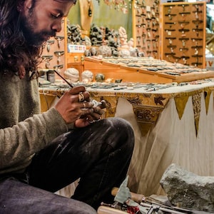 May include: A person working on a small stone sculpture with a brush and tools. The sculpture is on a table covered with a white tablecloth with a gold and brown patterned border. There are various tools and pieces of stone on the table.