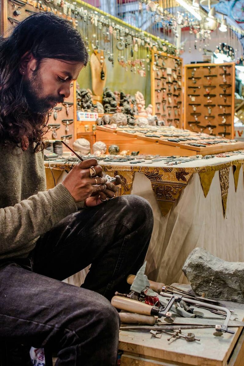 May include: A person working on a small stone sculpture with a brush and tools. The sculpture is on a table covered with a white tablecloth with a gold and brown patterned border. There are various tools and pieces of stone on the table.