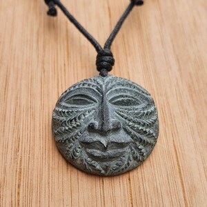 May include: A black cord necklace with a round, green stone pendant featuring a carved face with intricate details.