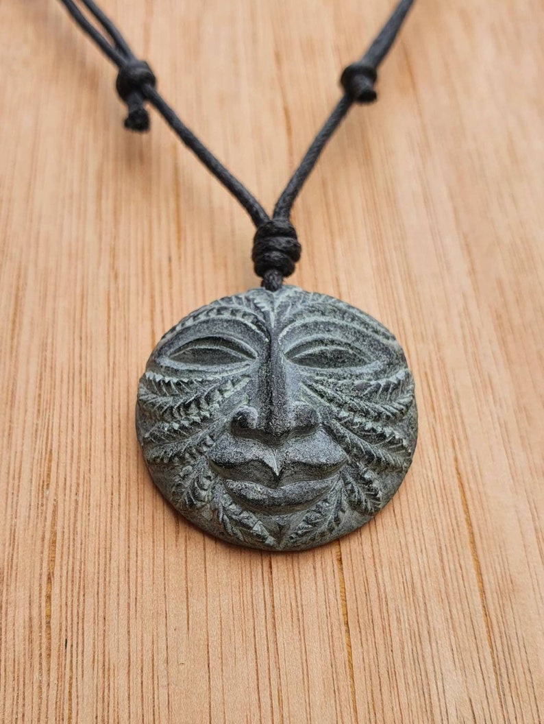 May include: A black cord necklace with a round, green stone pendant featuring a carved face with intricate details.