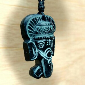 May include: Black pendant necklace featuring a carved wooden figure of a face with intricate details. The figure has a crown-like headpiece and a detailed face with eyes, nose, and mouth.