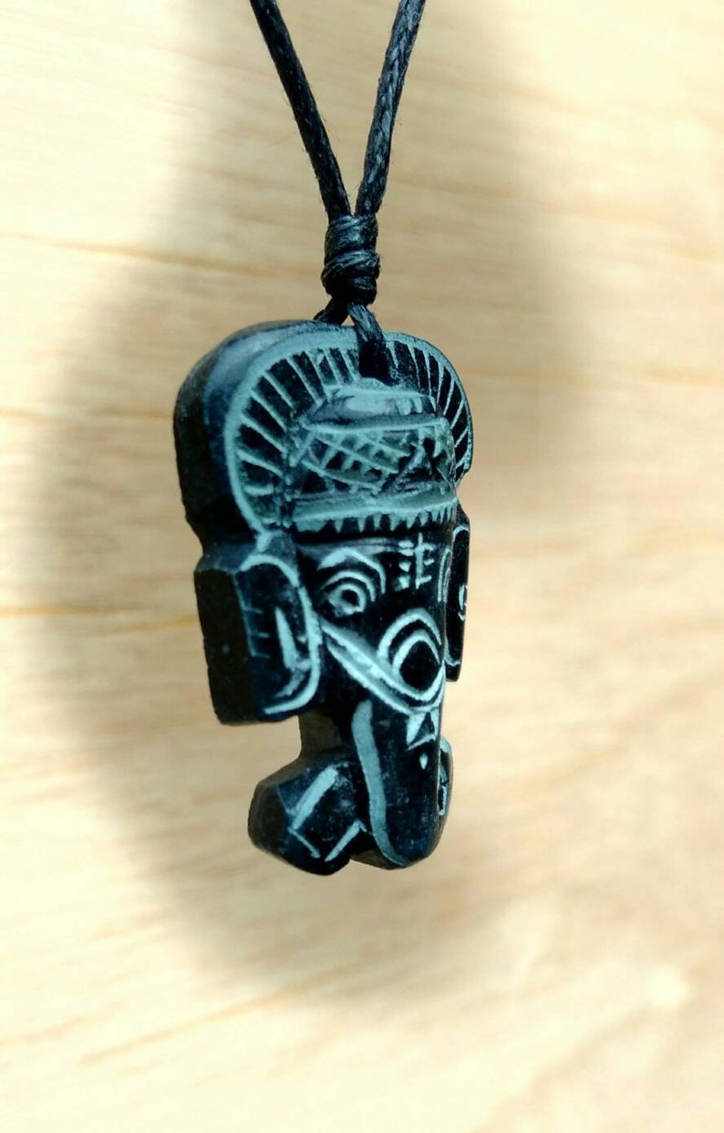 May include: Black pendant necklace featuring a carved wooden figure of a face with intricate details. The figure has a crown-like headpiece and a detailed face with eyes, nose, and mouth.