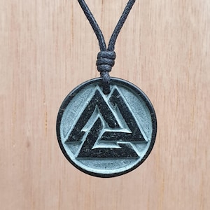 May include: A black pendant necklace featuring a circular, weathered-look charm with a stylized triangular symbol. The pendant is suspended from a black cord, creating a minimalist and rustic aesthetic. The background is a light brown wooden surface.