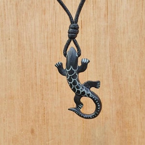Può includere: Una collana con ciondolo a forma di lucertola nera con un cordino nero annodato. La lucertola ha un design dettagliato con accenti bianchi sulla schiena e sulle zampe. Lo sfondo è una superficie di legno marrone chiaro.