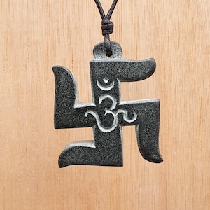 Puede incluir: Un collar con un colgante negro en forma de esvástica con la palabra "Om" inscrita en su interior. El colgante está hecho de un material oscuro y texturizado.