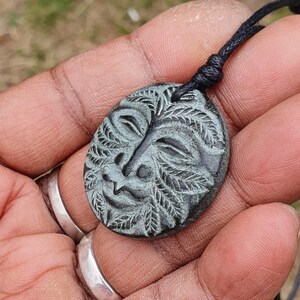 以下が含まれることがあります： 黒い紐にぶら下がった、葉で囲まれた彫刻された顔を持つ濃い緑色の石のペンダント