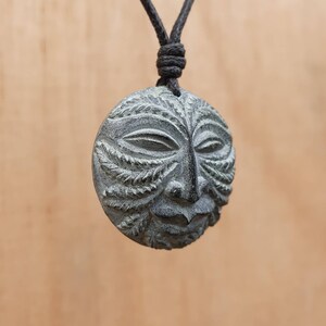 May include: A gray stone pendant necklace with a carved face design. The face has a leafy pattern around the eyes and mouth.