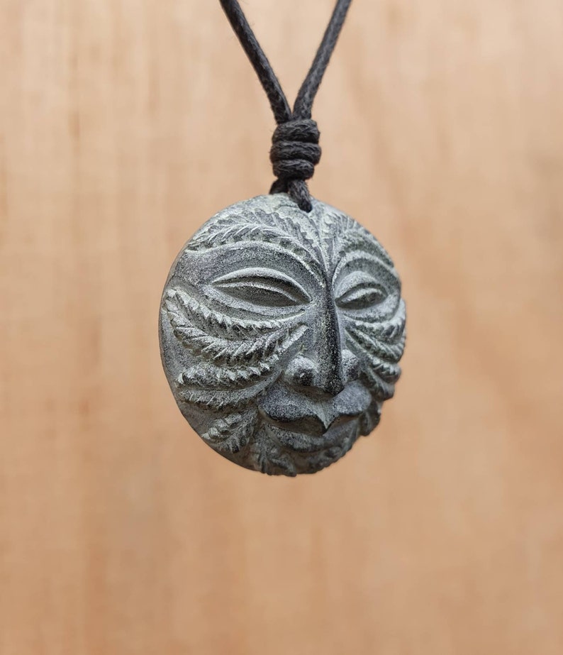 May include: A gray stone pendant necklace with a carved face design. The face has a leafy pattern around the eyes and mouth.