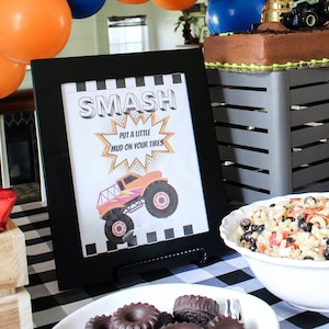 May include: A Monster Truck themed party table setting with a black and white checkered tablecloth. There is a framed sign that says "Smash Put a little mud on your tires" with a monster truck image. There is a bowl of pasta salad, a plate of chocolate tire shaped cookies, and a trophy with a monster truck on top.