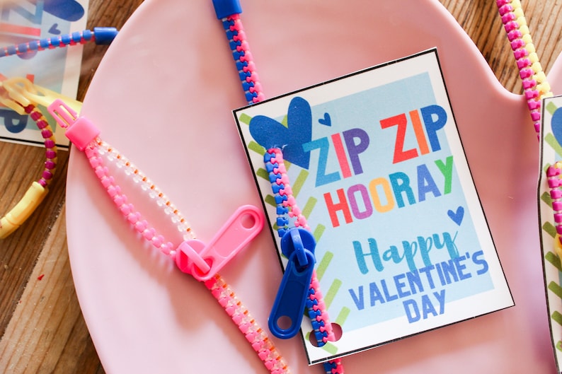 May include: A close-up of Valentine's Day themed zipper bracelets and a card. The card reads "ZIP ZIP HOORAY Happy VALENTINE'S DAY" in colorful text. The bracelets are pink, blue, and yellow with matching zipper pulls. The background is a pink surface.