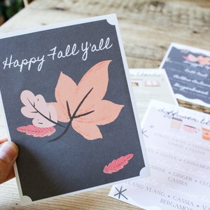 Può includere: Un biglietto con sfondo grigio scuro e il testo "Happy Fall Y'all" in scrittura bianca. Il biglietto presenta un'illustrazione stilizzata di un ramo con tre foglie arancioni e rosa.