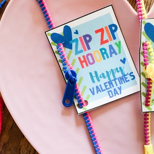 May include: A pink heart-shaped plate holds a Valentine's Day card with a blue zipper. The card reads "ZIP ZIP HOORAY Happy VALENTINE'S DAY" in colorful text. The zipper is blue and pink, with a blue pull. Other zippers are visible.