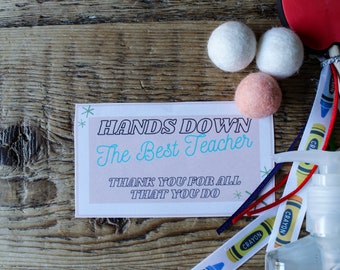 May include: A teacher appreciation gift set with a card that reads "Hands Down The Best Teacher Thank You For All That You Do", a bottle of hand sanitizer, a ribbon with crayon designs, and white and pink pom poms.