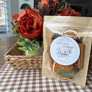 May include: A clear plastic pouch containing a simmer pot kit with dried orange slices, cinnamon sticks, star anise, and a bay leaf. The label features a circular design with the text The Cozy Cottage and a small house illustration.
