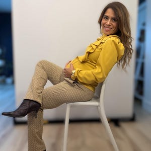 May include: A woman wearing a mustard yellow blouse with ruffled detailing and patterned trousers. The trousers have a geometric design. She is wearing brown heeled boots and a gold watch. She is sitting on a white chair.