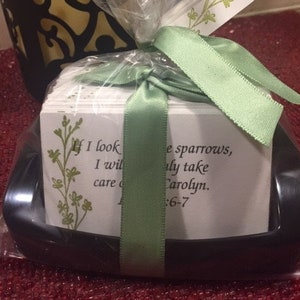 May include: A stack of small white cards with inspirational quotes and a green ribbon tied around them. The cards are wrapped in clear plastic and placed in a black tray. The text on the cards reads "If I look I will care" and "The sparrows, only take Carolyn. 6-7".