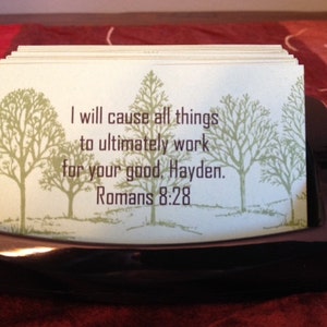 May include: A black frame holds a stack of cards with a green background and a message that reads "I will cause all things to ultimately work for your good. Hayden. Romans 8:28".