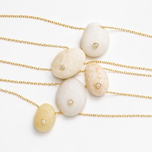 May include: Five necklaces with gold chains and pendant stones. Each stone is a different shade of white, cream, and yellow, and has a small, round, clear gem set in the center. The necklaces are displayed on a white background.