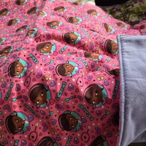 May include: A pink blanket featuring a repeating pattern of a cartoon character, stethoscopes, and hearts. The blanket has a soft, light blue border. The design includes the text "I Care!"