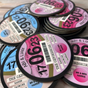 May include: A stack of round, black and white paper coasters with a design that resembles a car tax disc. The coasters feature a gold circle with the text "The Oxfordshire" and the year "2023". The text "Abi & Liam" is printed on the blue and pink coasters.