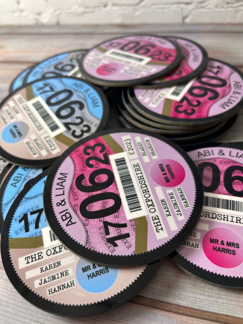 May include: A stack of round, black and white paper coasters with a design that resembles a car tax disc. The coasters feature a gold circle with the text "The Oxfordshire" and the year "2023". The text "Abi & Liam" is printed on the blue and pink coasters.