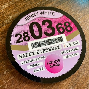 May include: A black and pink round sticker with the text "JENNY WHITE" and the date "28 03 68". The sticker also has the text "HAPPY BIRTHDAY" and "&pound;55.00". There are several other words on the sticker, including "CAMPING TRIPS", "BERYL", "FLUTE", "BEST FRIEND", "DANCING", and "I BELIEVE IN PIGS".