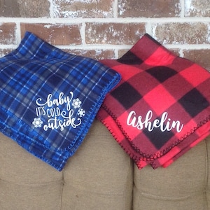 May include: Two fleece blankets, one blue plaid with "baby it's cold outside" and snowflakes, the other red and black buffalo plaid with the name "Ashelin". Both blankets are folded and displayed on a neutral-colored couch.