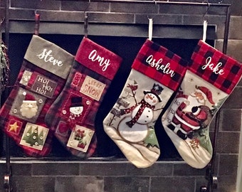 May include: Four Christmas stockings hang from a fireplace. The stockings are red, black, and white, with names embroidered on them. Each stocking has a different design, including a snowman, Santa Claus, and Christmas trees.