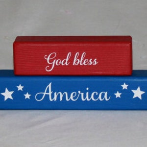 Puede incluir: Dos bloques de madera apilados uno encima del otro. El bloque superior es rojo y dice "God bless" en letras blancas. El bloque inferior es azul y dice "America" en letras blancas con estrellas blancas a cada lado de la palabra.