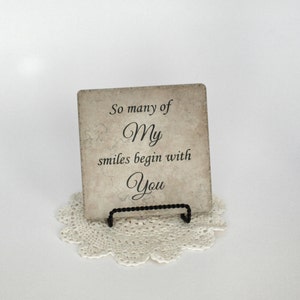 May include: A decorative tile with a beige background and black text that reads "So many of My smiles begin with You". The tile is on a white doily and a black metal stand.