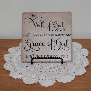 May include: A beige square tile with black text that reads "The Will of God will never take you where the Grace of God will not protect you". The tile is on a white doily.