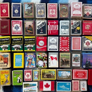 Può includere: Una collezione di cinquantacinque mazzi di carte da gioco assortiti disposti in una griglia. Le scatole rettangolari presentano vari design, tra cui loghi di marchi, paesaggi, bandiere nazionali e personaggi dei cartoni animati.