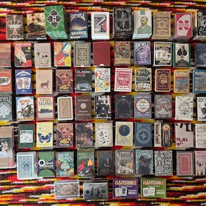 Puede incluir: Una gran colección de varias barajas de cartas dispuestas en una cuadrícula. Las barajas presentan diseños diversos, incluyendo temas como trenes antiguos, animales, cultura pop y la marca Bicycle, con muchas cajas que muestran ilustraciones únicas.