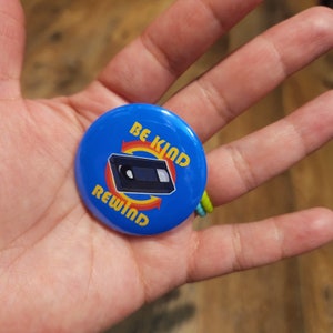 May include: A round, blue button with the words "BE KIND REWIND" in yellow, surrounding a graphic of a VHS tape. The button is held in a person's hand. The button is a retro-themed accessory.