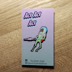 May include: A green alien with a large head and a ray gun is featured on this enamel pin. The pin is on a pink background with the words "Ack Ack Ack" in a bubble font.